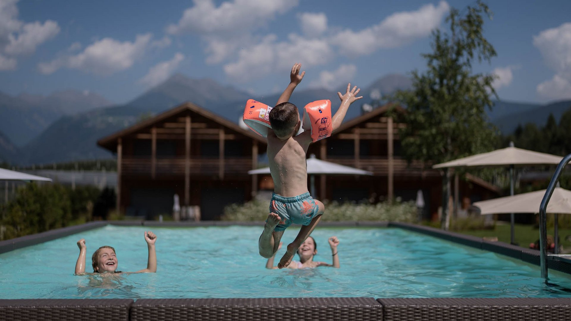 Kind mit Schwimmflügeln springt in Pool, zwei Kinder im Wasser, Bergkulisse