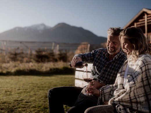 Holiday in Natz-Schabs? Kessler’s Mountain Lodge! Couple sitting outdoors by wooden house, man pouring red wine into glass