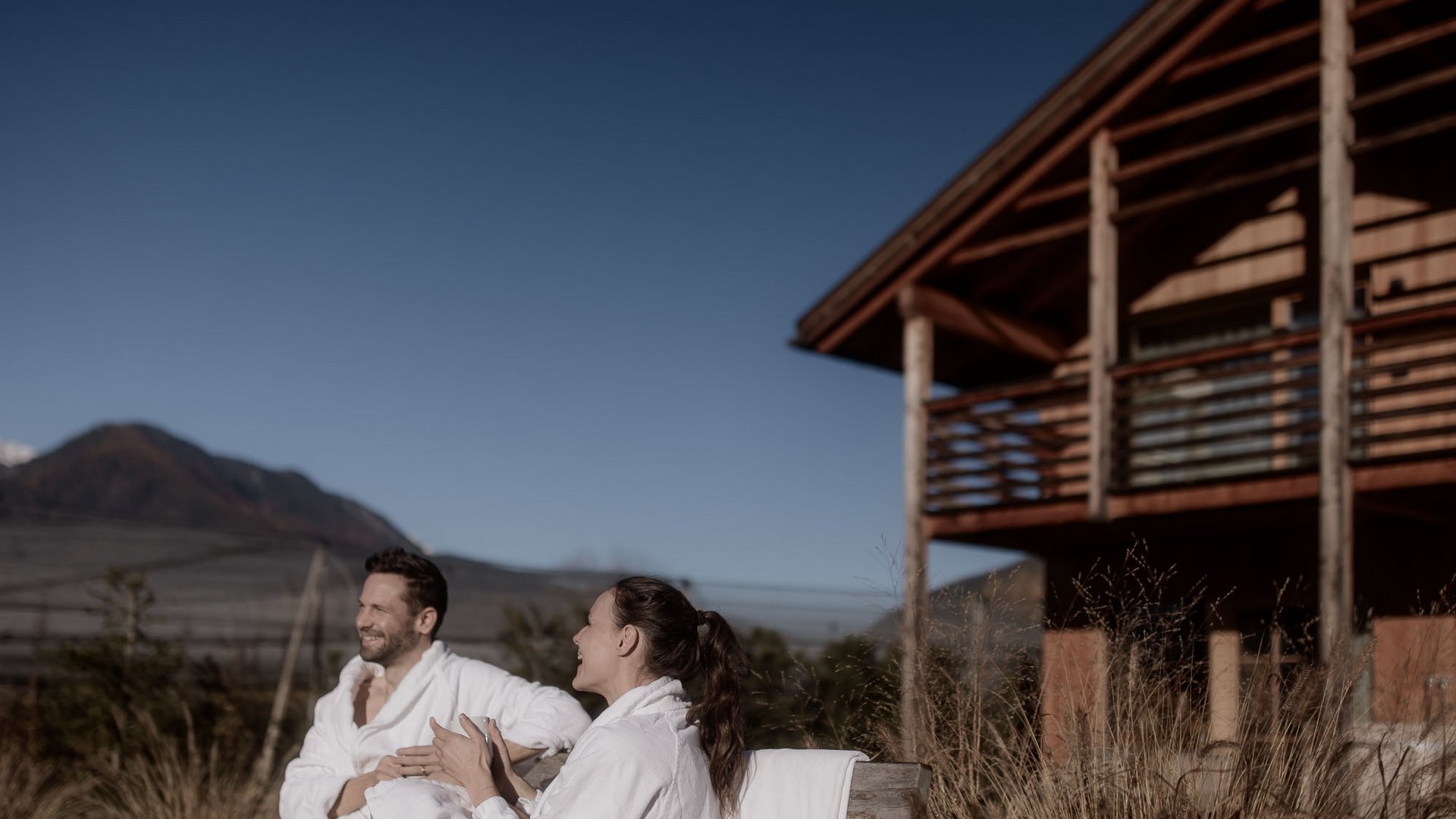 Appartamenti in Alto Adige: fuori dal comune Coppia in accappatoi rilassata davanti a una casa in legno sulle montagne