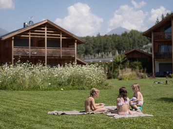 Für Familien Drei Kinder sitzen auf einer Decke auf einer Wiese vor Holzhäusern