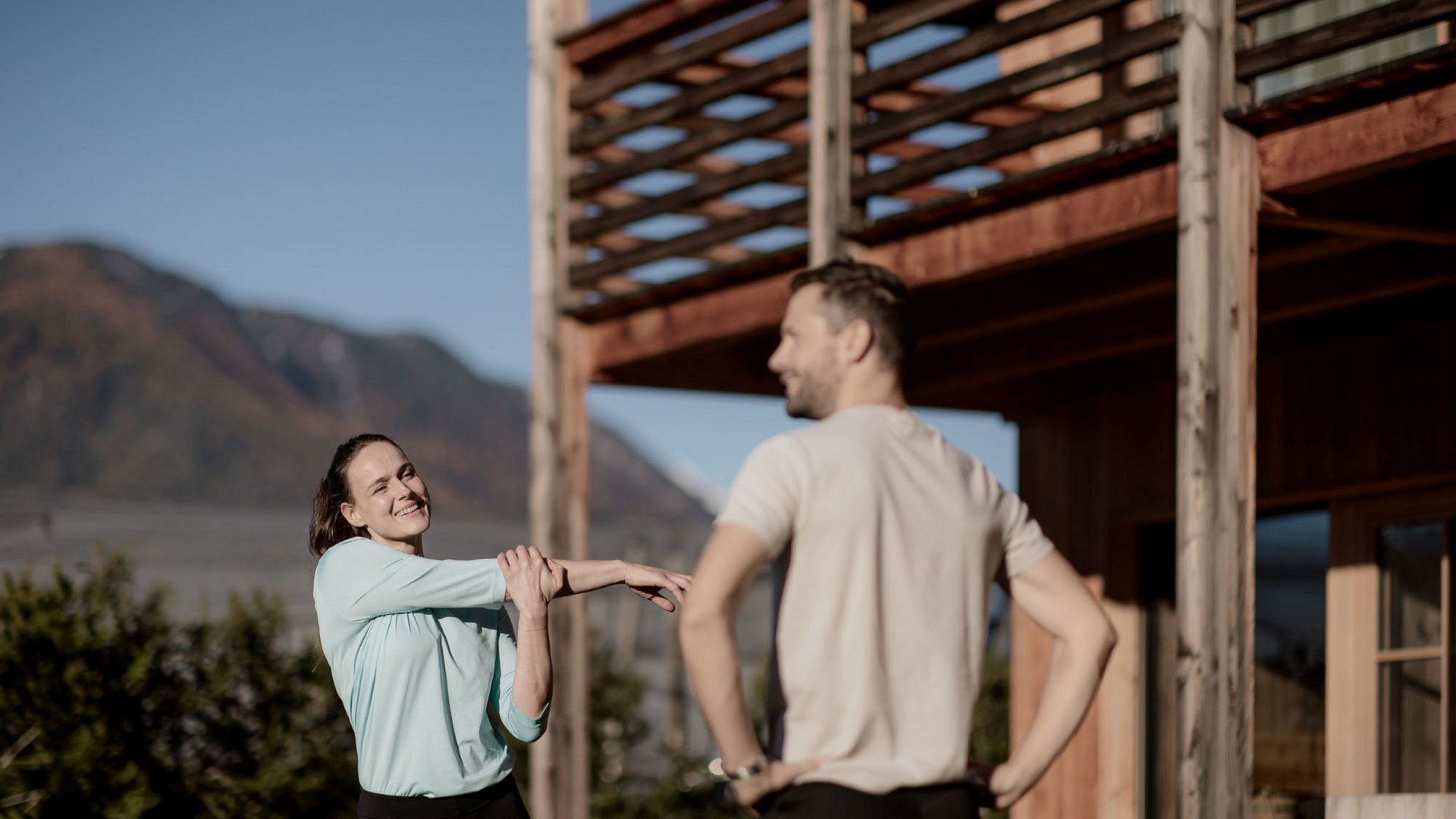 Urlaub in Natz-Schabs? Kessler’s Mountain Lodge! Frau dehnt Arm draußen, Mann schaut zu, Berg und Hütte im Hintergrund