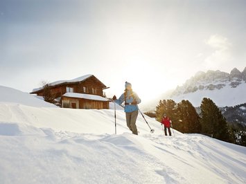 Agriturismo a Bressanone: vacanze invernali Due persone che fanno escursioni con racchette da neve davanti a una baita in montagna