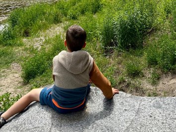 Appartamenti in Alto Adige: fuori dal comune Bambino seduto sulla riva del fiume con vista su montagne verdi e chiesa