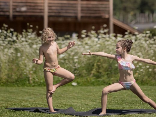 Für Familien Zwei Kinder machen Yoga auf der Wiese vor einer Holzhütte bei sonnigem Wetter