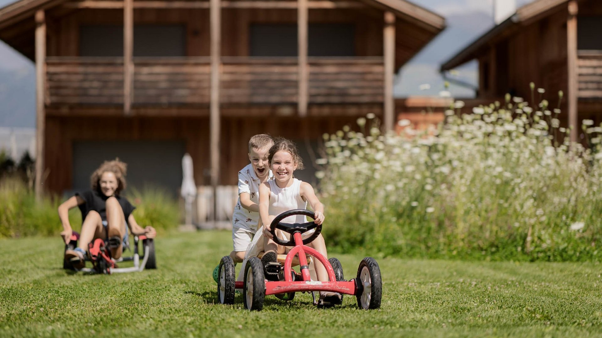 Für Familien Kinder fahren mit Tretfahrzeugen auf einer Wiese vor Holzhäusern