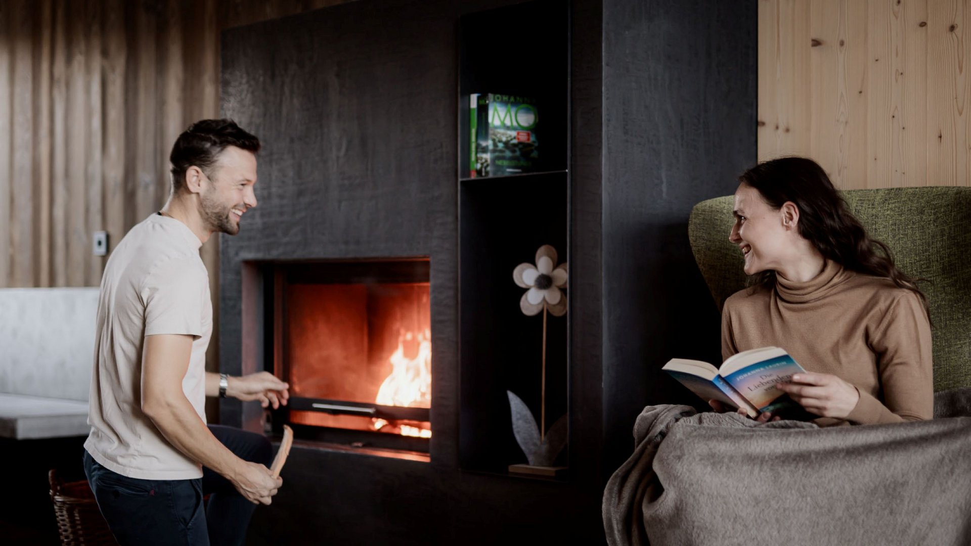 Urlaub in Natz-Schabs? Kessler’s Mountain Lodge! Paar entspannt sich am Kamin, Frau liest Buch, Mann fügt Holz hinzu