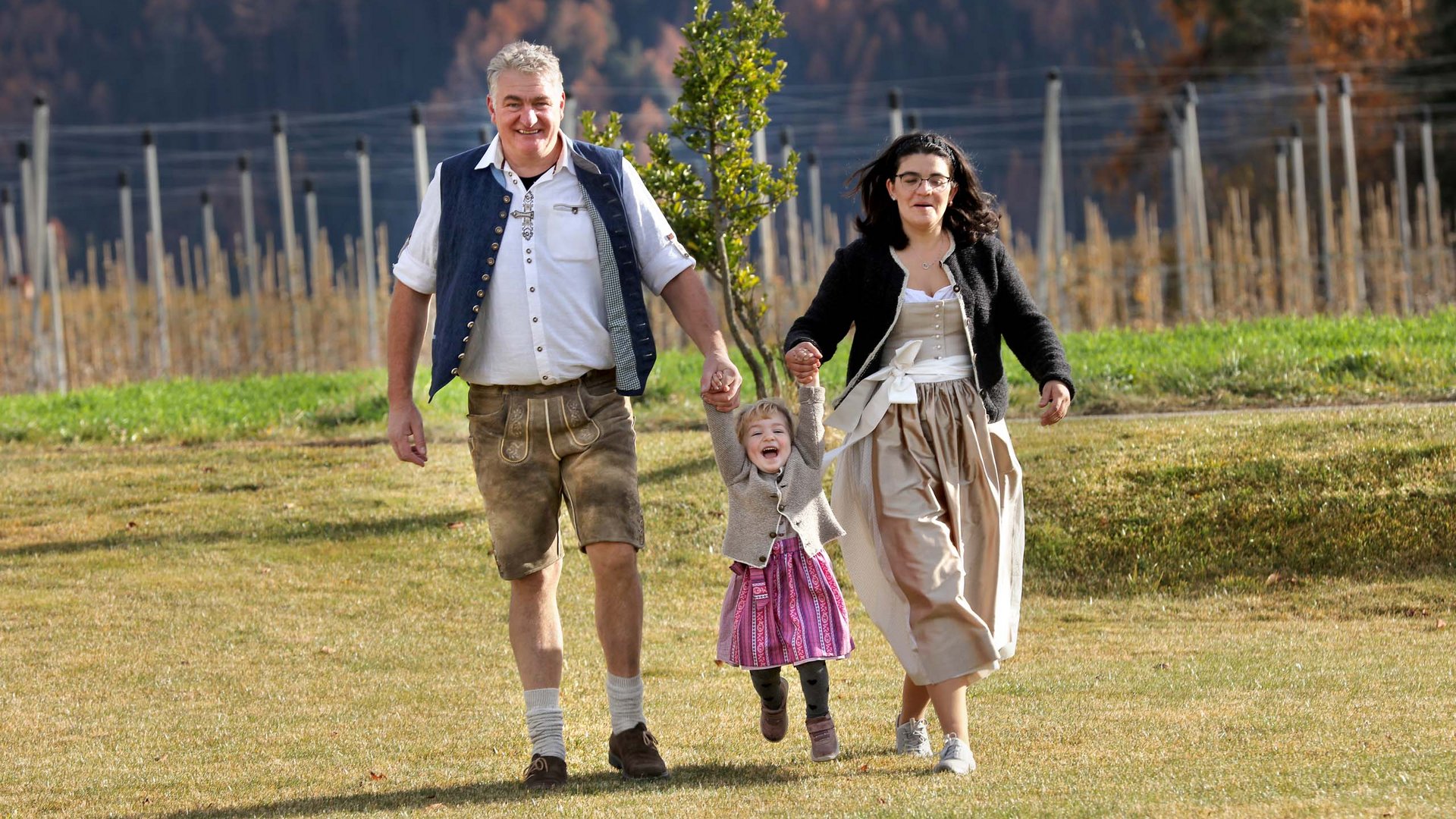 Vacanze in fattoria? Kessler’s Mountain Lodge! Famiglia felice in abbigliamento tradizionale cammina all'aperto tenendo il bambino per mano