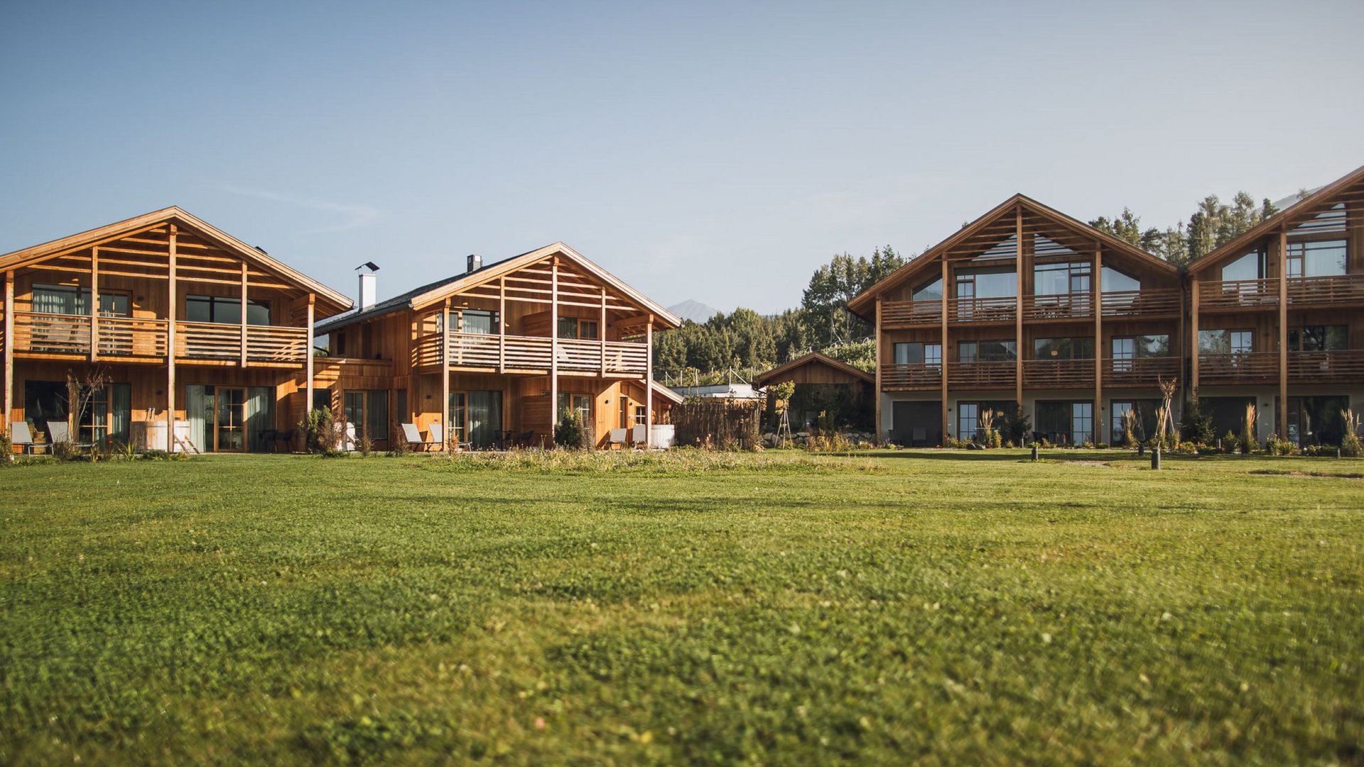 Urlaub in Natz-Schabs? Kessler’s Mountain Lodge! Holzhäuser mit Balkonen vor großem grünem Rasen und klar blauem Himmel