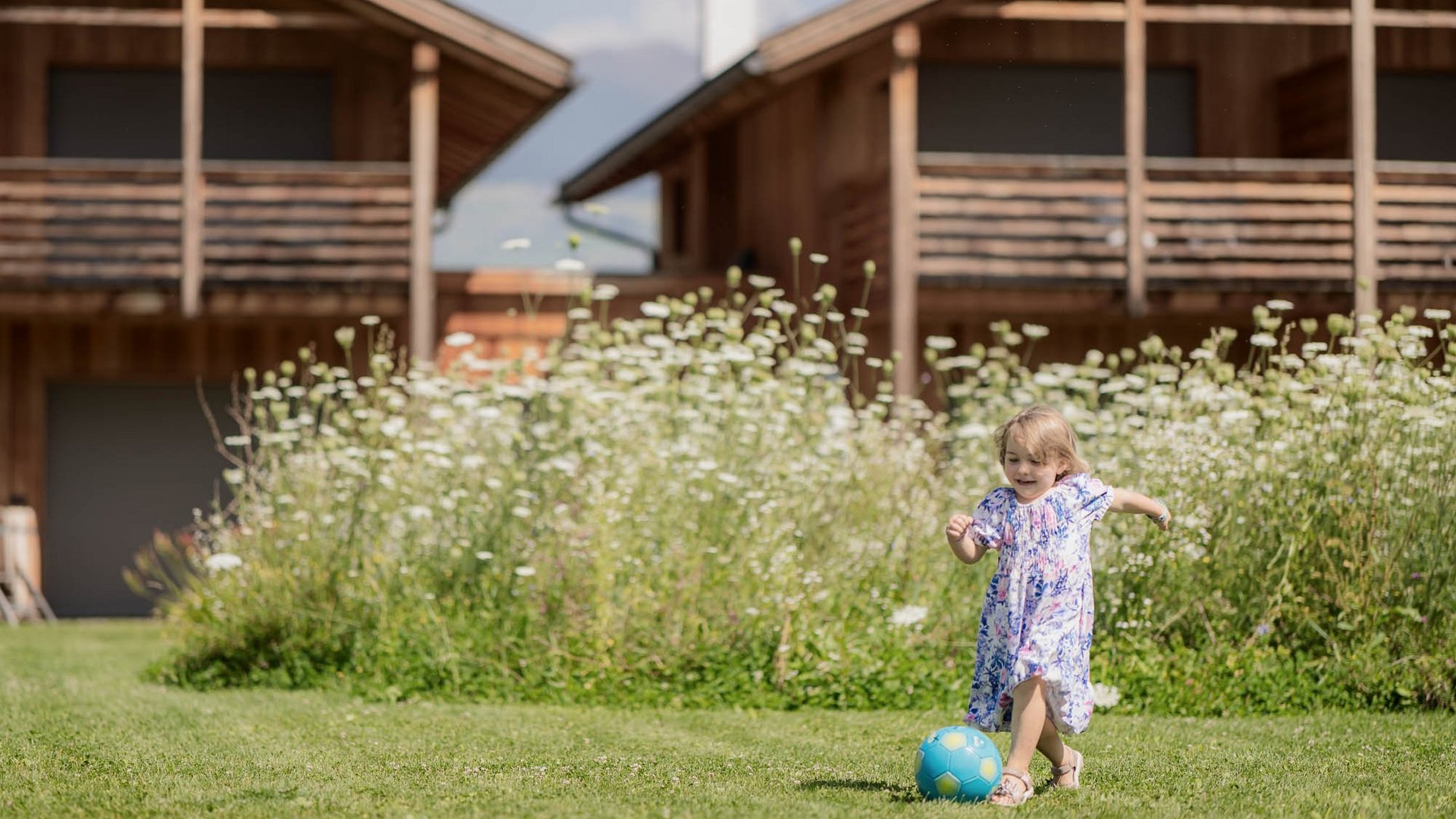 Für Familien Kleines Mädchen spielt mit blauem Ball auf grünem Rasen vor Holzhäusern