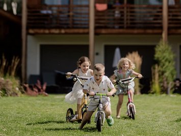 Für Familien Drei Kinder fahren auf Rollern vor einem Holzhaus auf dem Rasen