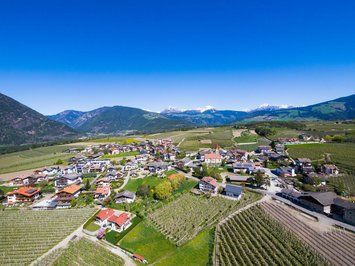 Urlaub mit Hund in Südtirol: Kessler’s! Luftaufnahme eines kleinen Dorfes umgeben von Feldern und Bergen unter klarem Himmel