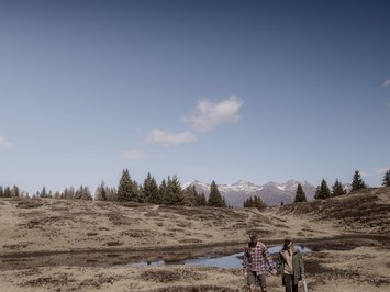 Holiday with a dog in South Tyrol: Kessler’s! Couple hiking on mountain trail with pine trees and snowy mountains in the background.