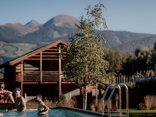 Urlaub in Natz-Schabs? Kessler’s Mountain Lodge! Gäste entspannen im Pool vor Holzhäusern und Bergkulisse