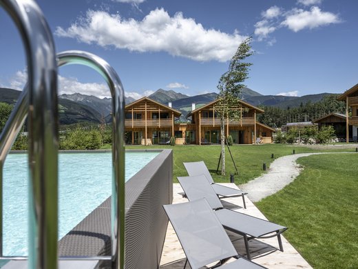 Urlaub in Natz-Schabs? Kessler’s Mountain Lodge! Außenpool mit Liegestühlen vor Holzhäusern und Bergen im Hintergrund