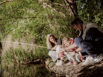 Für Familien Famiglia con bambino che pesca vicino a un fiume sotto alberi verdi